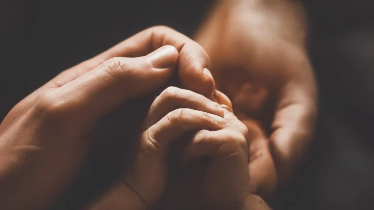 Una mano adulta sosteniendo suavemente la mano de un bebé, rodeadas por una cálida luz dorada.
